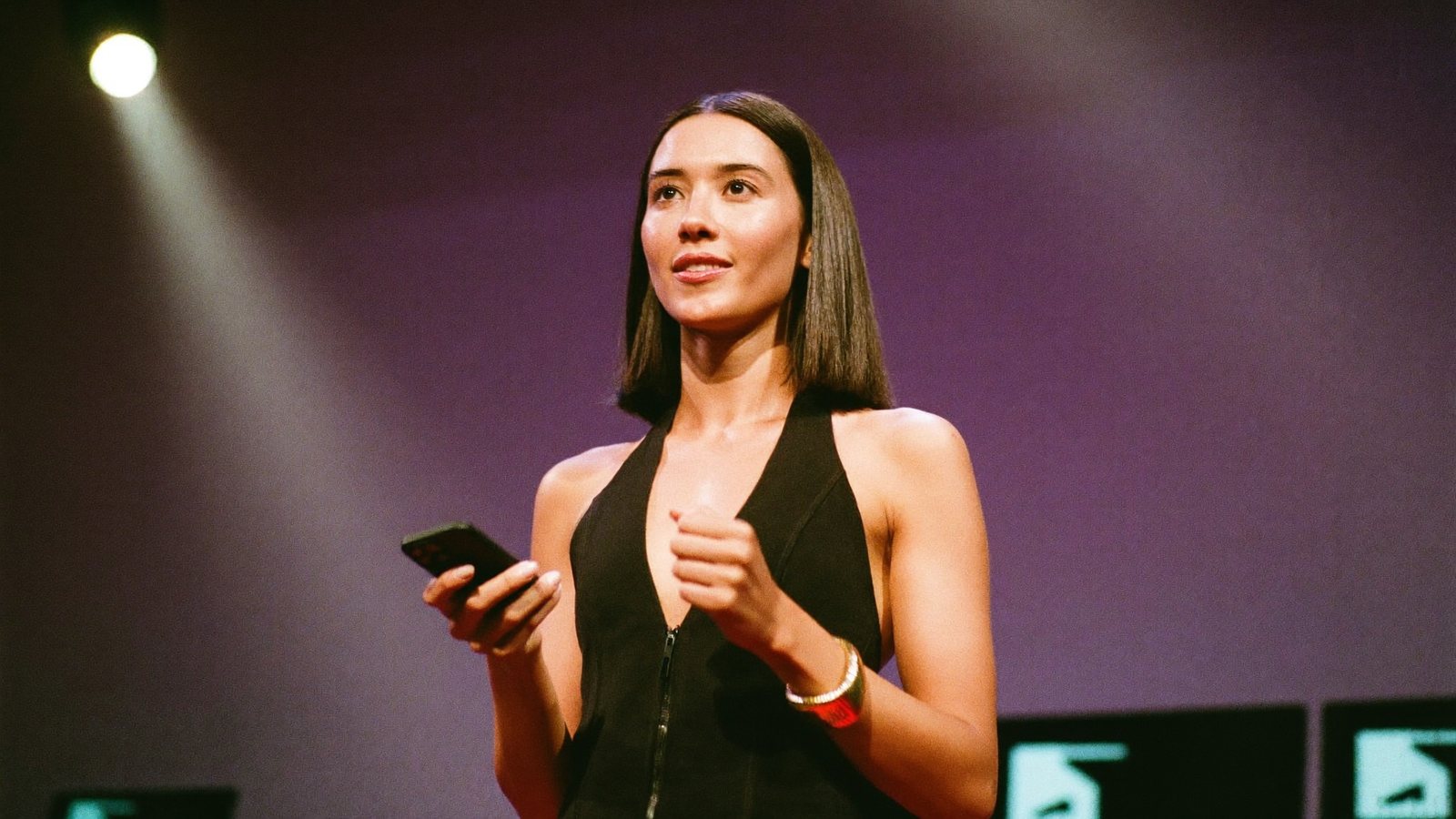 A speaker mid-sentence on a small stage under a spotlight, a phone on a tripod in the front row filming her