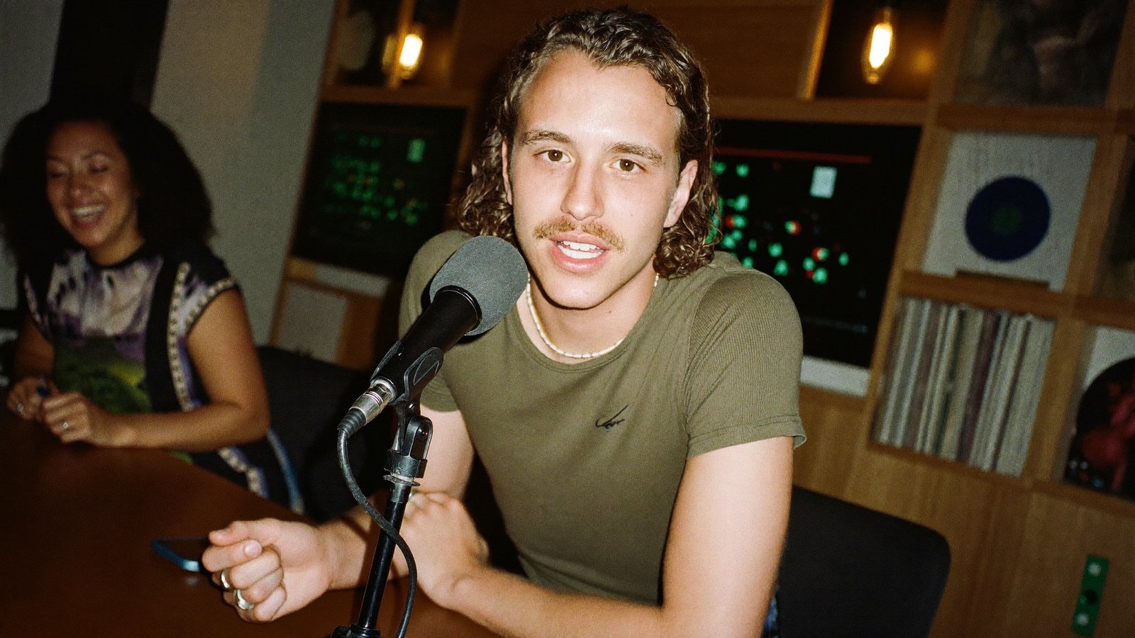 A podcast host leaning into a broadcast mic across the table from a laughing guest, a phone clamped to the boom filming vertically
