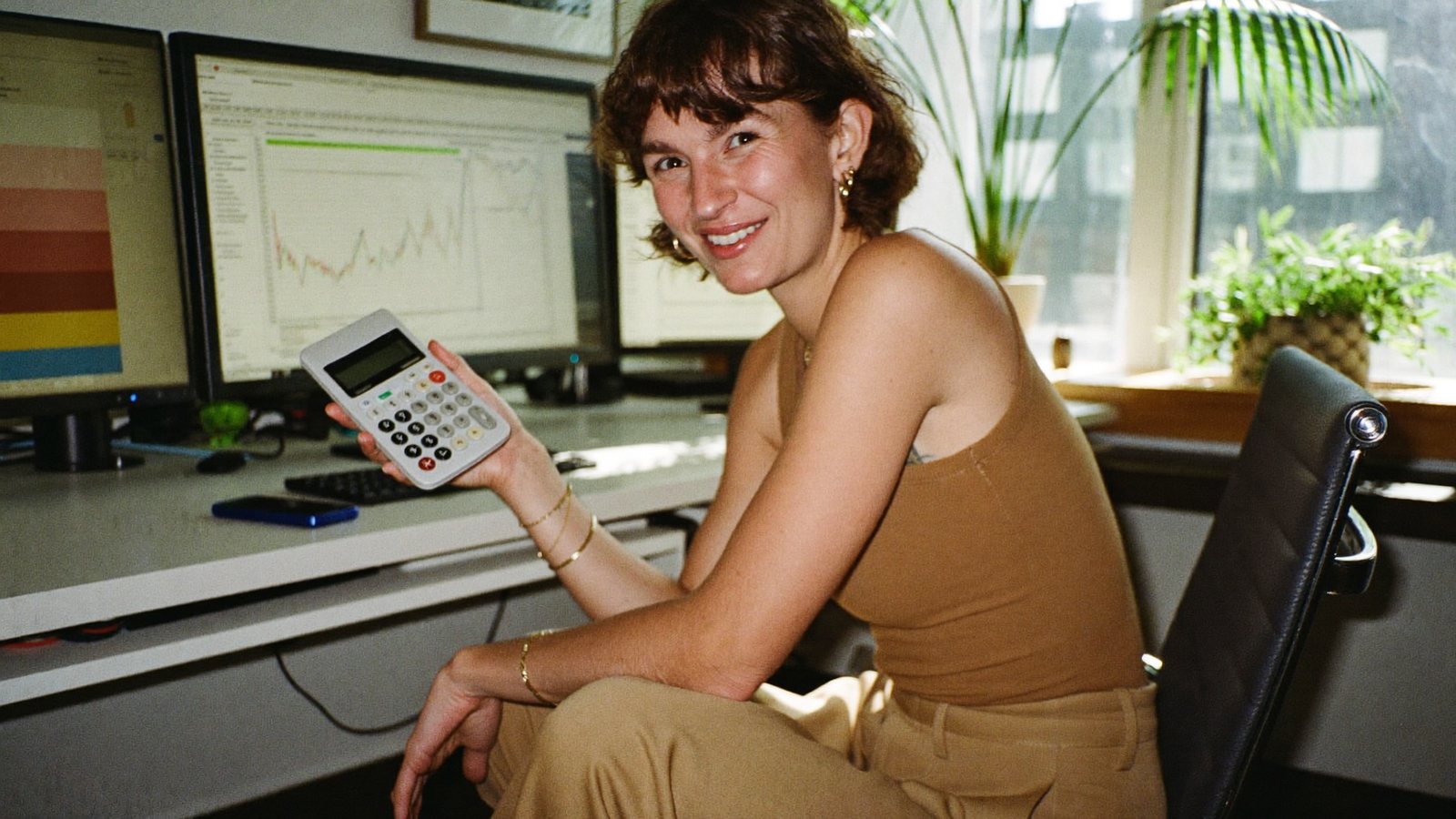 A CPA at a clean desk with a single monitor, recording herself on a phone on a tripod while holding up a calculator with a laugh