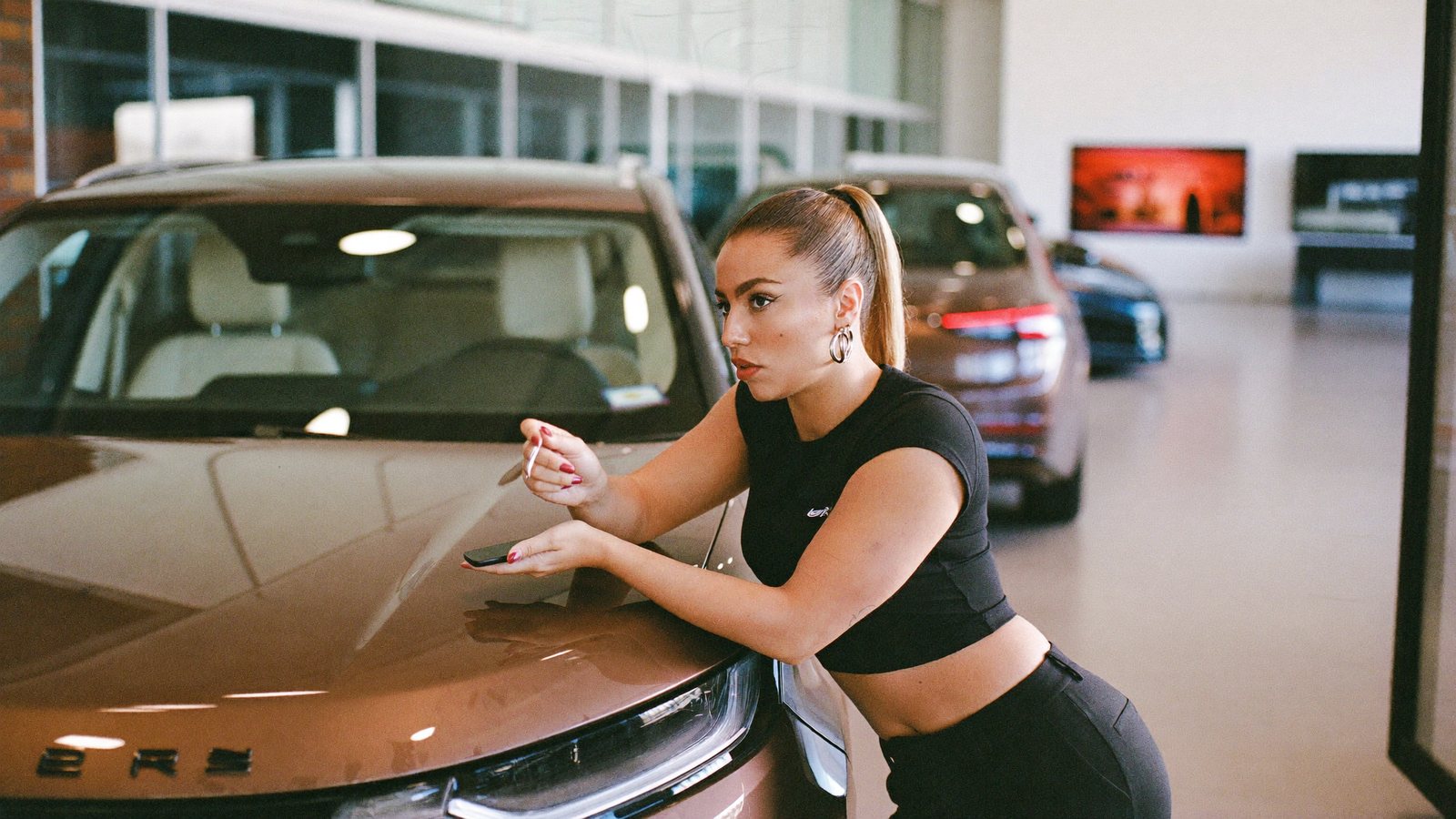 A dealership sales lead recording a vertical video on a phone beside an SUV on a bright showroom floor