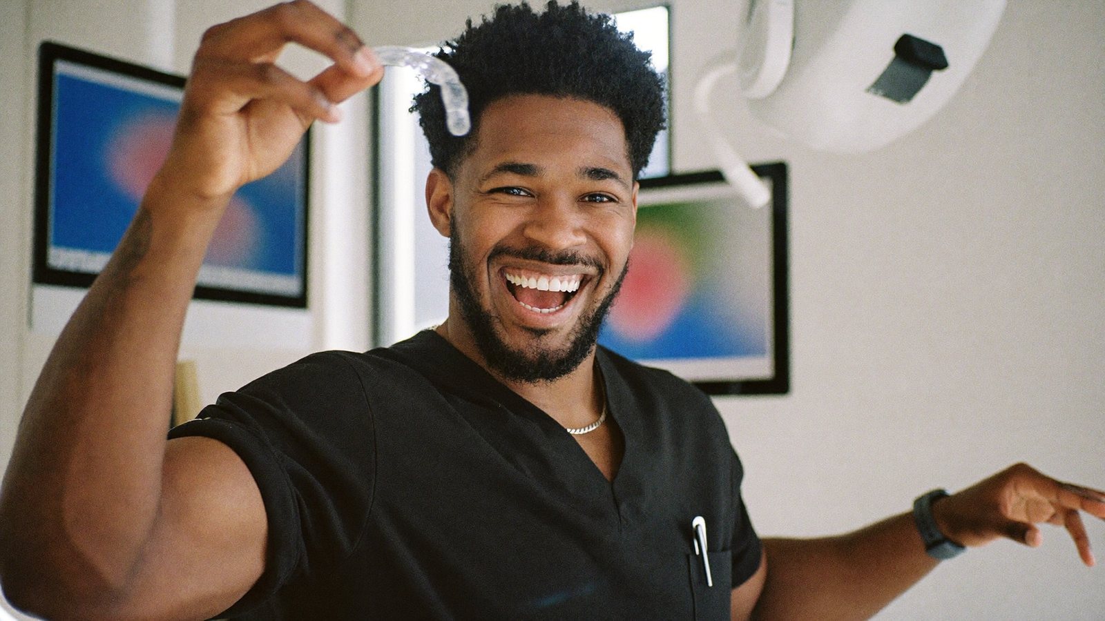 A dentist in black scrubs in a sleek dental suite, recording himself on a phone on a tripod while laughing and holding a clear aligner up to camera