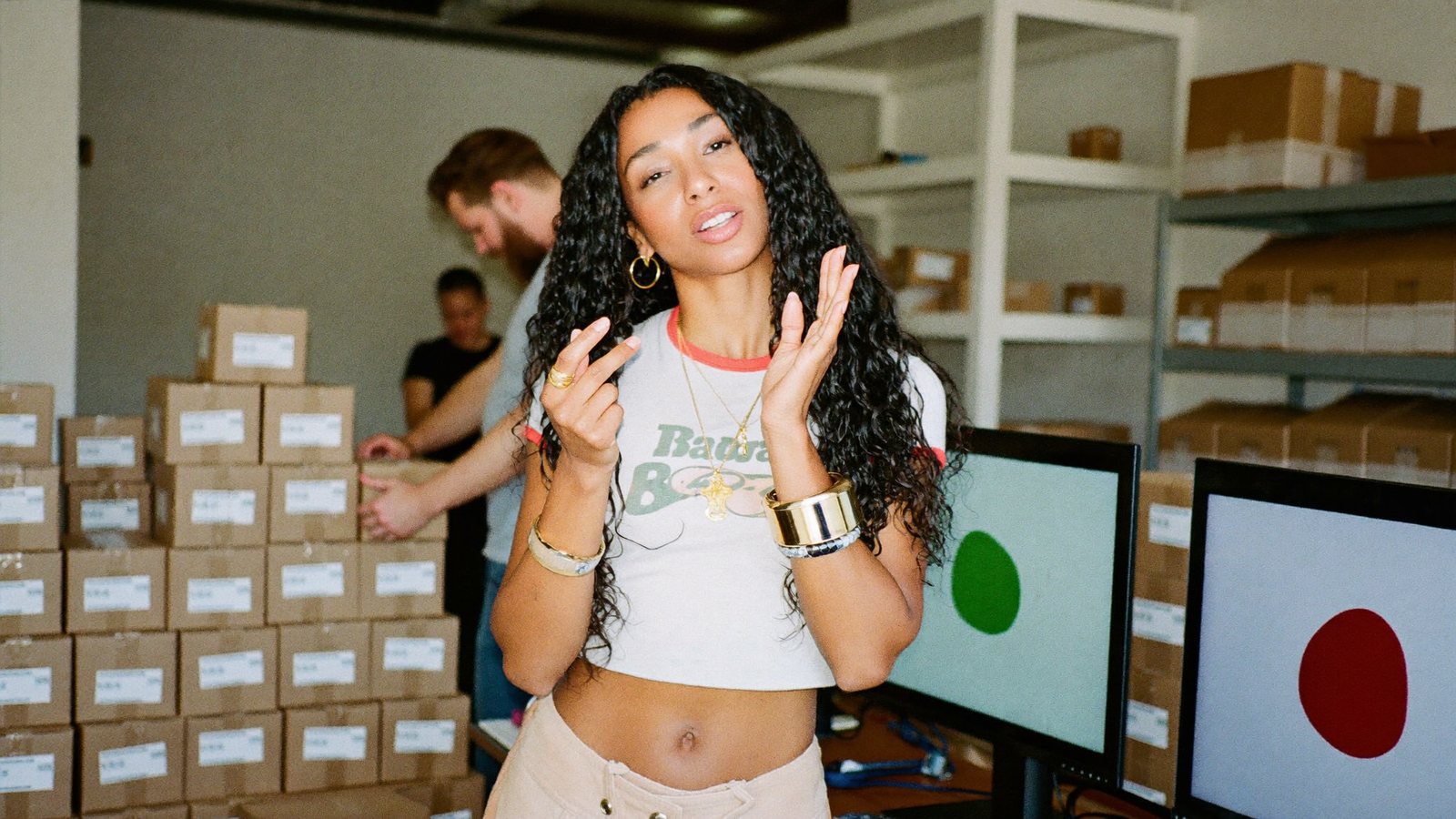 A DTC brand founder in a packing room stacked with mailers, holding a product up to a phone on a ring-light tripod with a big grin