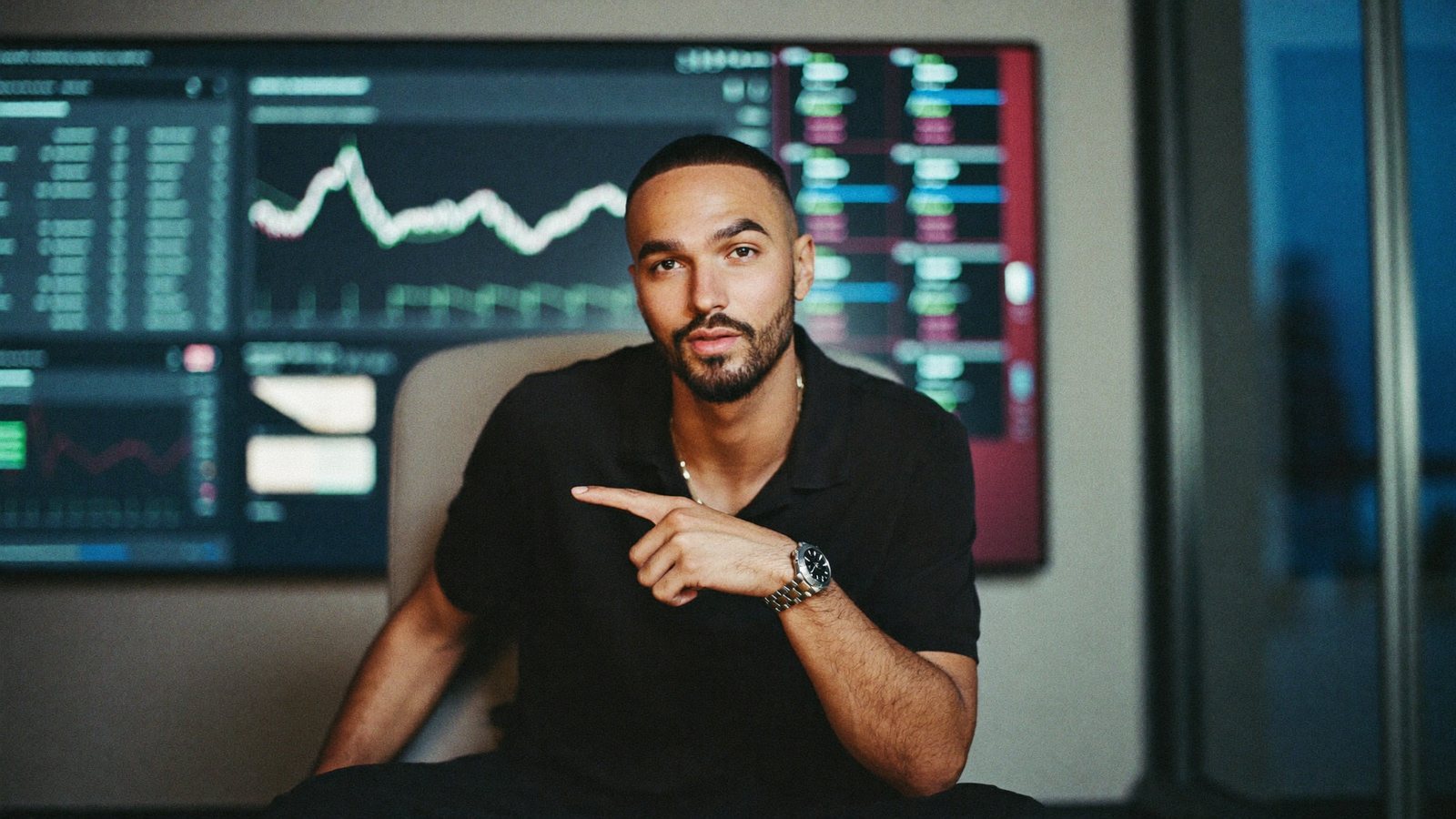 A fintech product lead in a modern office with a wall-sized dashboard of soft charts, recording himself on a phone on a tripod and pointing at a number