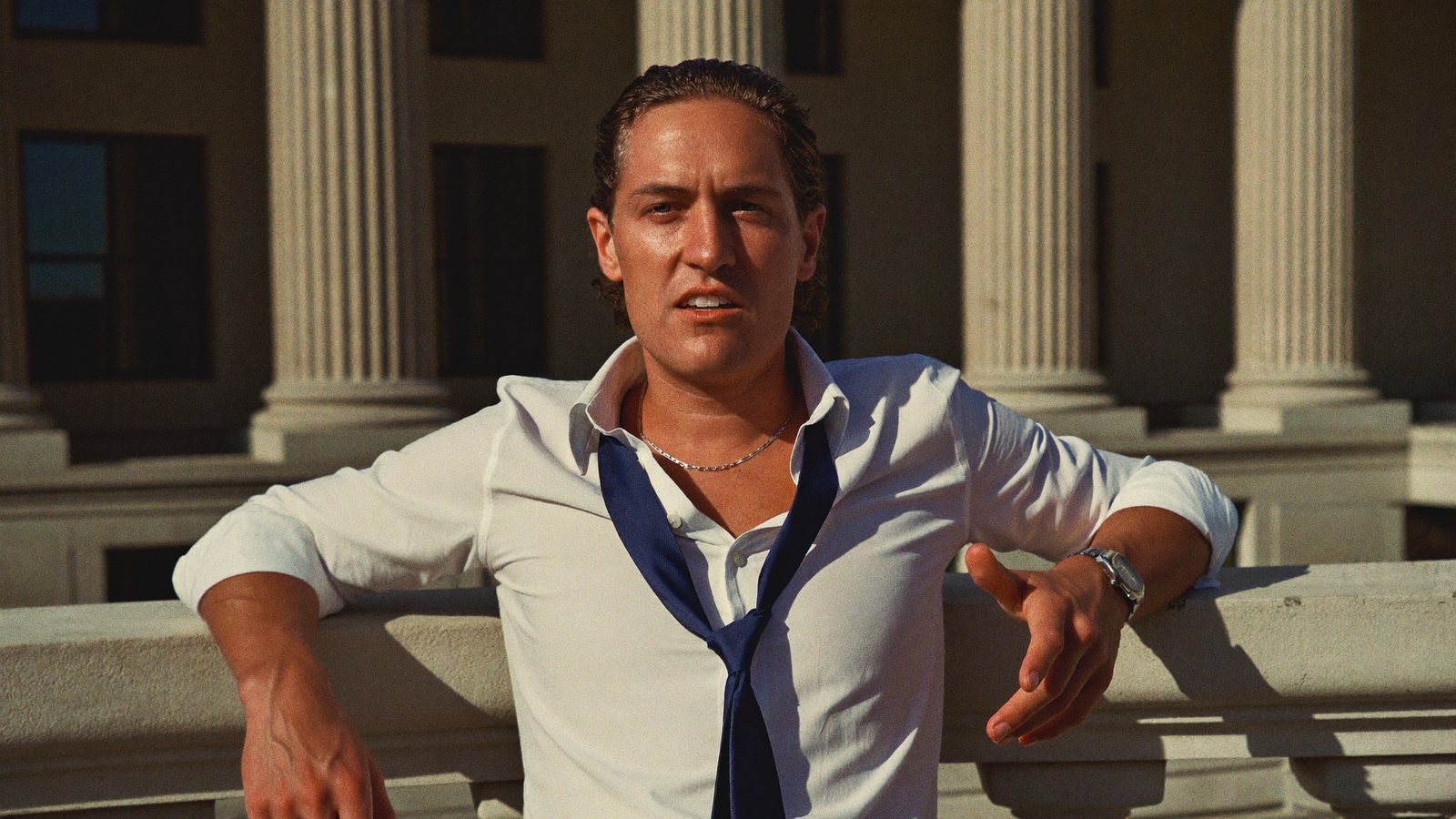 A litigation attorney leaning on a marble courthouse railing with columns behind, recording himself on a phone on a mini tripod and pointing to camera