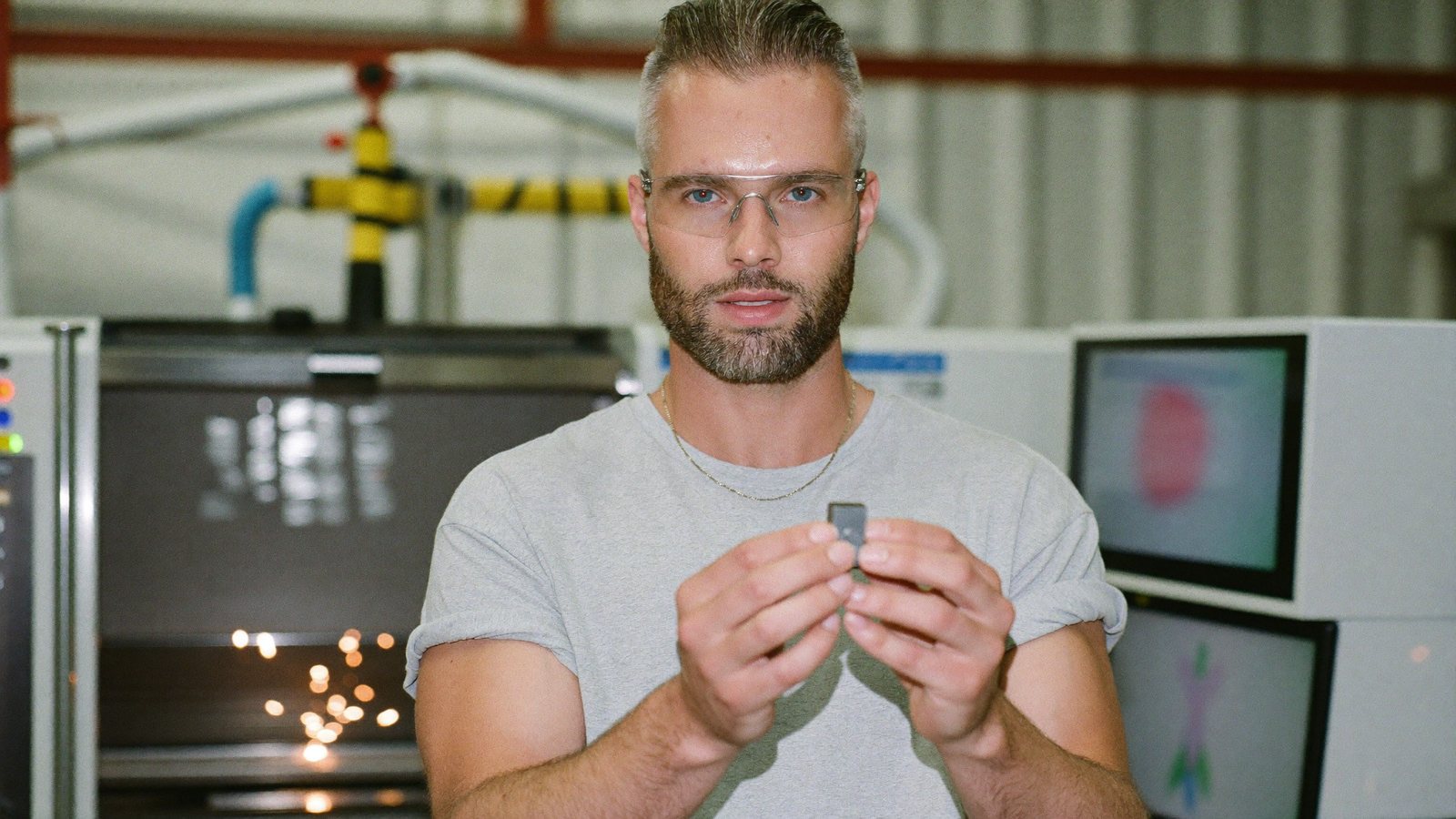 A manufacturing company owner on a bright factory floor beside a CNC machine, holding up a machined part to a phone on a tripod