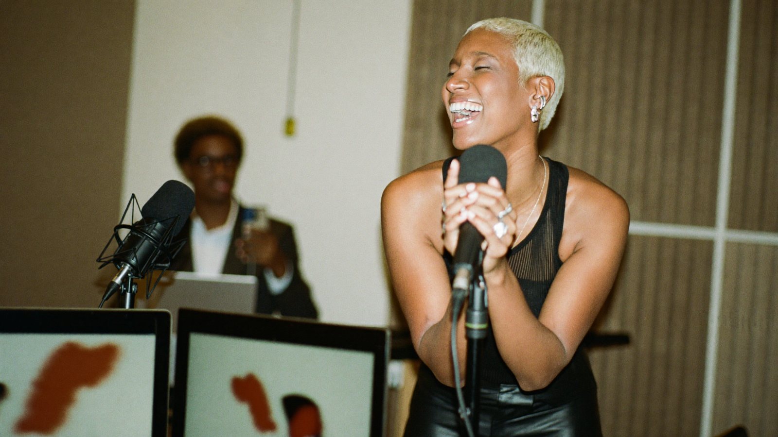 A media founder in a podcast studio leaning into a broadcast mic and laughing while a phone on a tripod films her