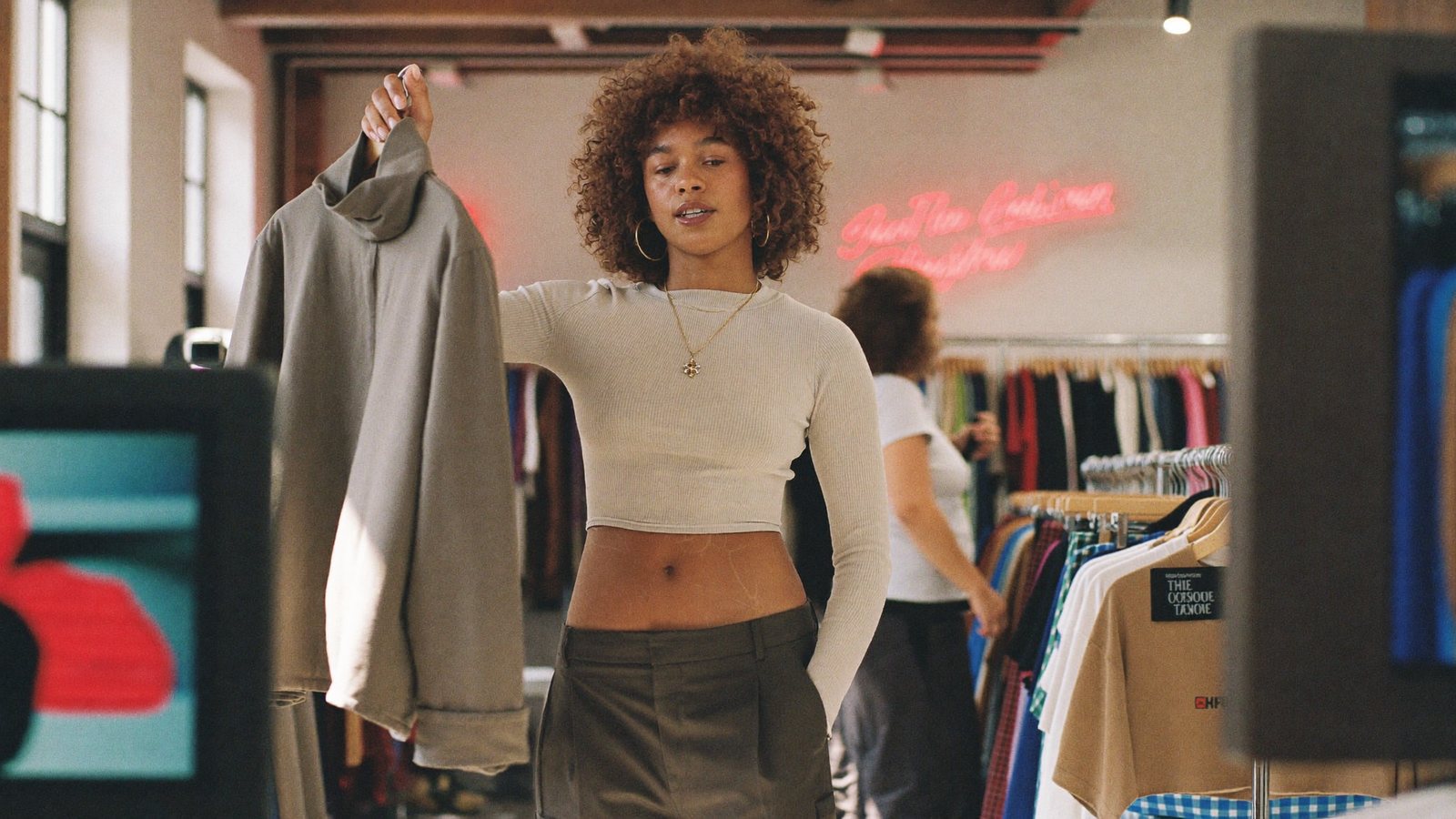 A boutique owner in a sunlit shop holding a jacket up to a phone on a ring-light tripod